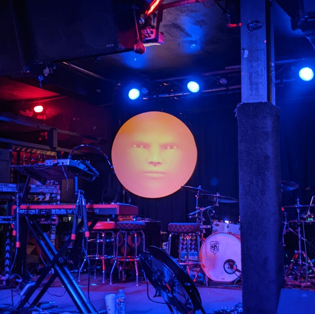 A picture of an empty stage at a TWRP show. Behind the band's instruments and gear, an expressionless orange face regards the camera.